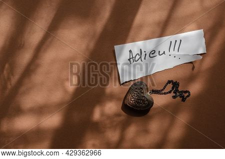 Handwritten Word Adieu In A White Piece Of Paper And A Wooden Amulet In The Shape Of A Heart. The Wo