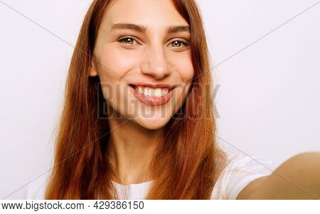 Attractive ginger female taking selfie with mobile phone. Beautiful young woman having cheerful expression while posing against white background.