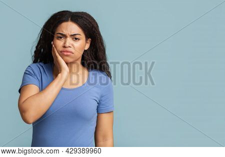 Aching Teeth. Unhappy Black Lady Suffering From Toothache Pain Touching Cheek Over Blue Background W