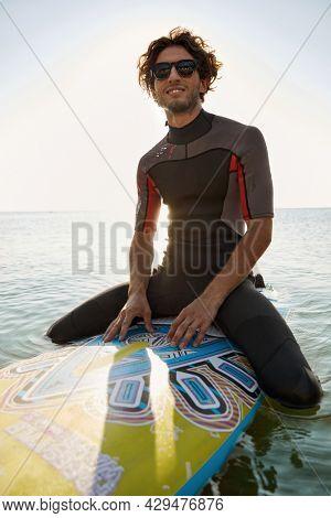 Young surfer sitting on surfboard in sea water. Enjoying curly man wear diving suit and glasses. Concept of extreme water sport. Idea of summer vacation. Sunny daytime