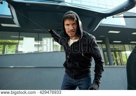 Aggressive european robber looking in car trunk. View from auto trunk with human victim. Male bearded bandit wear black hoodie and gloves. Concept of kidnapping. City daytime