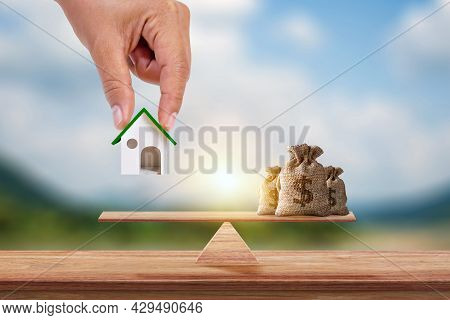 Hand Holding Model Of House And Savings Bag Placed On Scales And Blurred Green Background. Real Esta