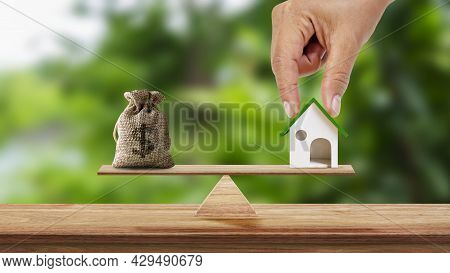 Hand Holding Model Of House And Savings Bag Placed On Scales And Blurred Green Background. Real Esta