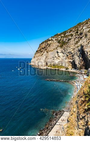 Sorrento, Italy - August 26 2020: Small Beach Full Of Colorful Sunbeds And Umbrellas Next To High Cl