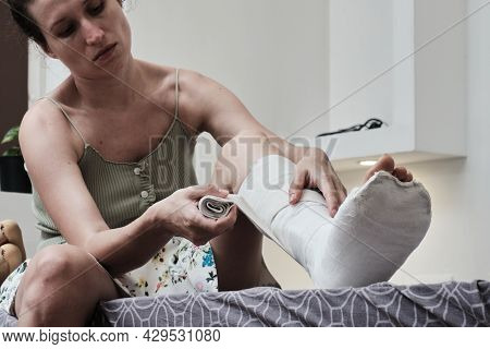 Woman Sitting On A Bed Puts An Elastic Bandage On A Broken Leg. The Leg Is In A Plaster Cast. Home R