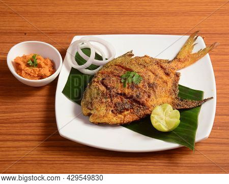 Pomfret Fish Tawa Fry Garnished With Lemon And Onion In A White Ceramic Plate With Wooden Background