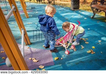 Little Cute Toddler Boy And A Beautiful Year-old Baby Girl In Blue Clothes Play In A Pink Toy Stroll