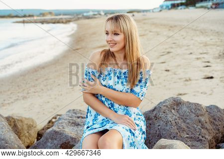 Young Blonde Woman In Summer Dress Sitting On The Rocks On The Beach And Enjoying The Moment. Caucas