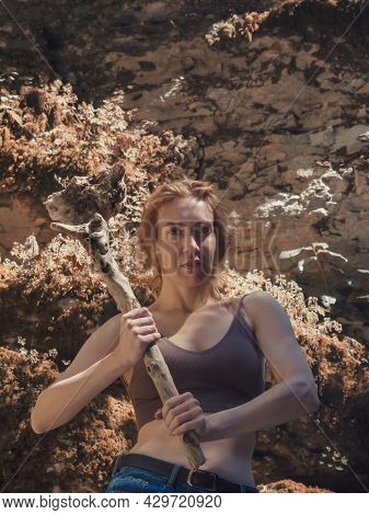 A Girl With A Serious Face Holds A Driftwood Club In Her Hands While Standing On Mossy Rocks Under T