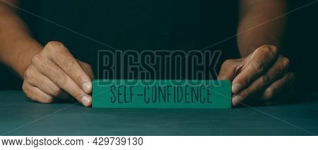 a caucasian man shows a green building block with the text self-confidence written in it, on a dark gray surface, in a panoramic format to use as web banner or header
