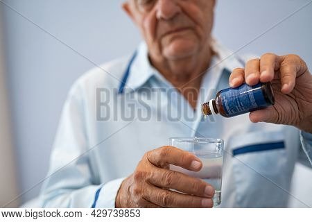Close up of wrinkled hands of senior man pouring melatonin drops in glass of water at clinic. Elderly male dripping medical liquid into cup of water at nursing home before sleeping, pouring medicine.
