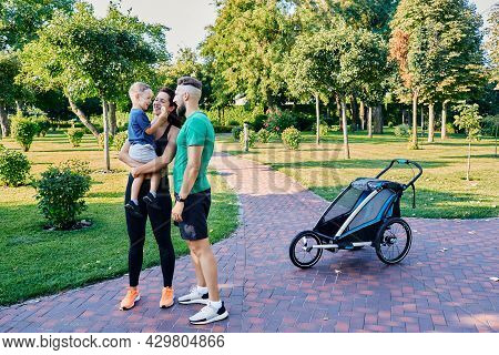 Active Family After Exercising And Running With Jogging Stroller At City Park. Happy Couple With Son