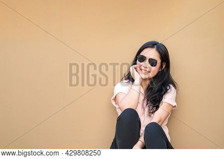 Relaxed Pretty Asian Woman Sits On The Rooftop Floor With A Light Brown Background And Smiles To The