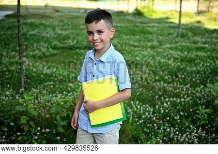 Happy Elementary Student Coming Back To School. Adorable Schoolboy Holding A Workbooks And Smiling T