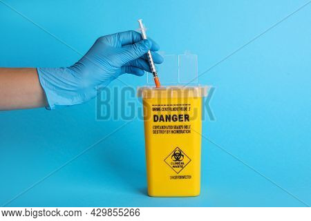 Doctor Throwing Used Syringe Into Sharps Container On Light Blue Background, Closeup