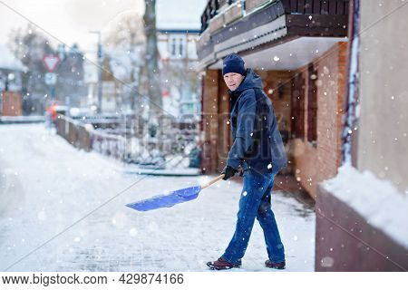 Man With Snow Shovel Cleans Sidewalks In Winter During Snowfall. Winter Time In Europe. Young Man In