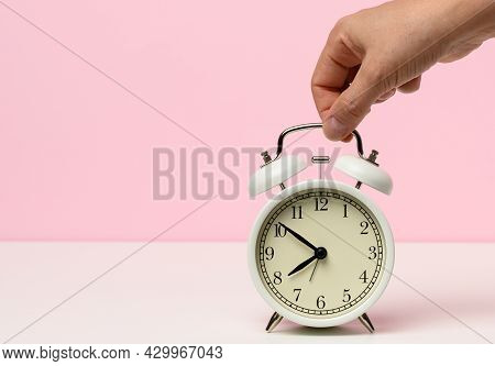 Female Hand Holds A White Metal Alarm Clock On A White Table, Time Is Ten To Eight In The Morning