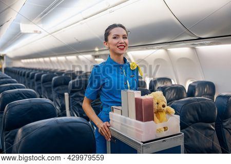 Cheerful Female Air Hostess In Bright Blue Uniform Smiling At Camera While Leading Trolley Cart With