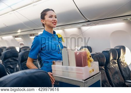 Pretty Stewardess In Bright Blue Uniform Smiling Aside While Leading Trolley Cart With Gifts Through
