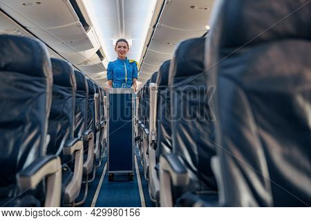 Female Cabin Attendant Leading Trolley Cart Through Empty Plane Aisle