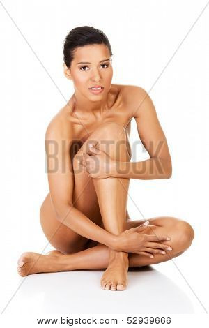 Beautiful naked young woman sitting. She is covering her nudity by her leg and hands. Isolated on white. 