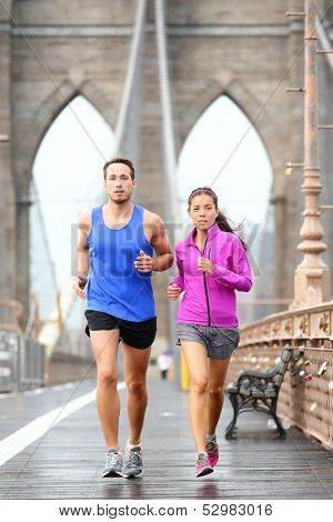 Running couple jogging training for New York marathon. Runners in rain outside. Asian woman and Caucasian man runner and fitness sport models jogging on Brooklyn Bridge, New York City, USA