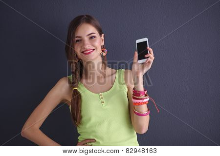 Beautiful young woman holding cellphone, standing near dark wall