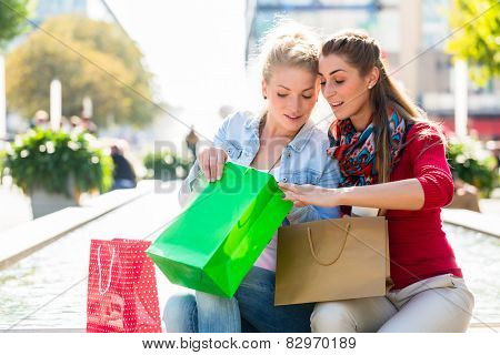 Two women friends sitting on fountain rim in city with shopping bags