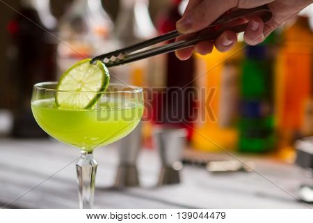 Green drink in coupe glass. Tongs hold slice of lime. Delicious beverage with sour juice. Fresh taste of kiwi daiquiri.