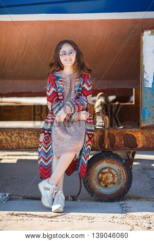Cute teen girl, caucasian appearance, brunette with long curly hair, wearing sunglasses, wearing a long coat knitted red and blue, spending time in the countryside in the fresh air in summer, standing near a rusty truck for transportation of boats