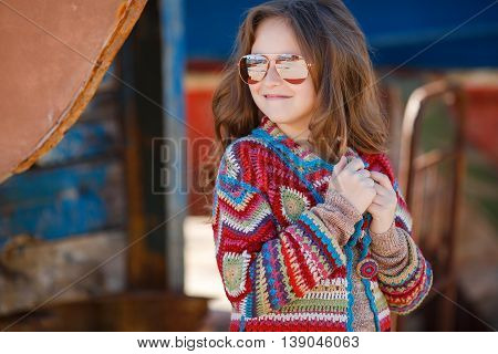 Cute teen girl, caucasian appearance, brunette with long curly hair, wearing sunglasses, wearing a long coat knitted red and blue, spending time in the countryside in the fresh air in summer, standing near a rusty truck for transportation of boats
