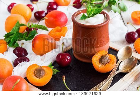 Food and drink, healthy nutrition concept. Fresh homemade yogurt in a ceramic pot with organic ripe apricots and cherries on a rustic wooden white table. Selective focus, summer breakfast composition