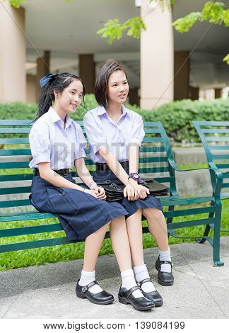 Cute Asian Thai high schoolgirls student couple in school uniform sit wih a happy smile face together on a bench