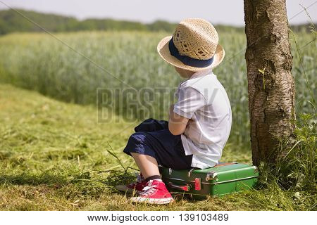 Cute little kid boy in shorts white polo and straw hat sits on his green suitcase in a field near a tree waiting for a bus. summer portrait of a small child in profile retro style. Ready to travel.