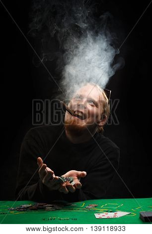 Beautiful young red-haired man with fuming cigar in shirt with long sleeves sitting at poker table and enjoys win holding chips in hands at black background.