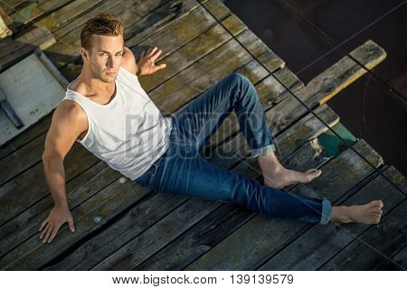 Cute barefoot guy lies on the wooden surface outdoors. He wears a white singlet and blue jeans and looks into the camera while leans on his hands. Photographed from the upper point. Horizontal.