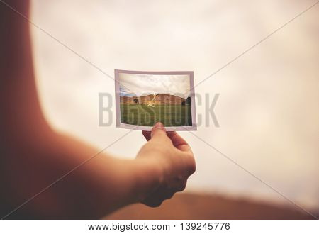 a woman holding an instant photo of a hill in front of her - destination or perseverance concept toned with a retro vintage instagrm filter app effect 