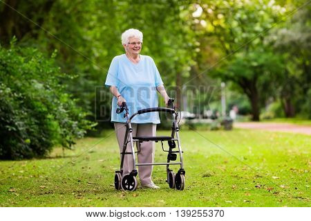 Happy senior handicapped lady with a walking disability enjoying a walk in a sunny park pushing her walker or wheel chair aid and support during retirement concept.