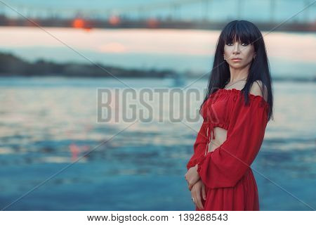 Portrait of a woman in a red dress on the river bank at sunset.