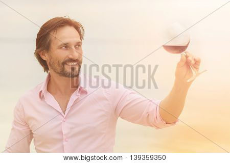 Toned picture of professional sommelier tasting red wine in glass. Happy smiling man is satisfied by taste of red wine isolated on sea.