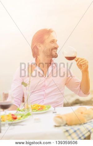 Toned picture of handsome man drinking red wine while sitting in restaurant or cafe by sea. Handsome bearded man in pink shirt relaxing and enjoying bunny day.