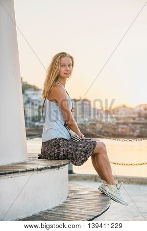Young blond tourist woman enjoying sunset and sitting on bench of the Lighthouse of Alanya by the sea. Meditarranean region, Turkey.