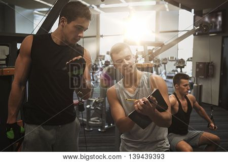 sport, fitness, lifestyle and people concept - men with clipboard taking notes and exercising on gym machine