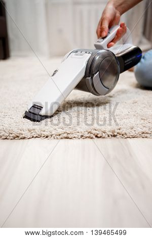 Female hand holding small white cordless vacuum cleaner and cleaning rug indoors