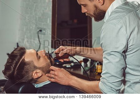 Hipster client visiting barber shop. The hands of young barber making the cut of beard