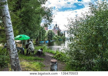 Pereslavl Zalessky, Russia - July 21, 2016: The unknown painter in leather boots and with a black cat on hemp draws an imperceptible tree on other river bank though directly before him a wonderful landscape.