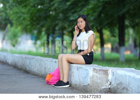 Girl female teenager with black hair in a bob white east Asian woman green park with a backpack background. Lady holds dials push your finger smiling looking into the camera talking on the phone happy.