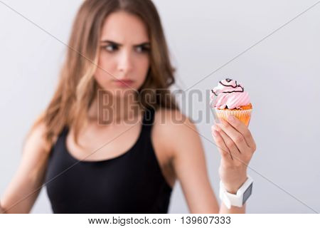 Resist the temptation. Pleasant baffled woman holding cupcake and thinking of eating it while standing isolated on grey background