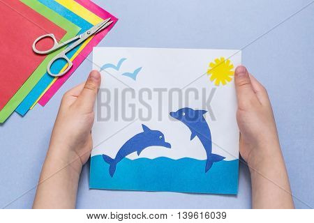 Paper applique is made by the child on a sea theme. The idea for children's creativity an art project made of paper. Sheets of colored paper glue scissors. Children's hands holding a picture