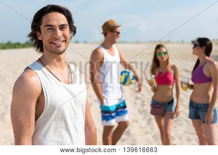 Nice company. Cheerful handsome man smiling and standing on the beach while resting with his friends who is standing in the background